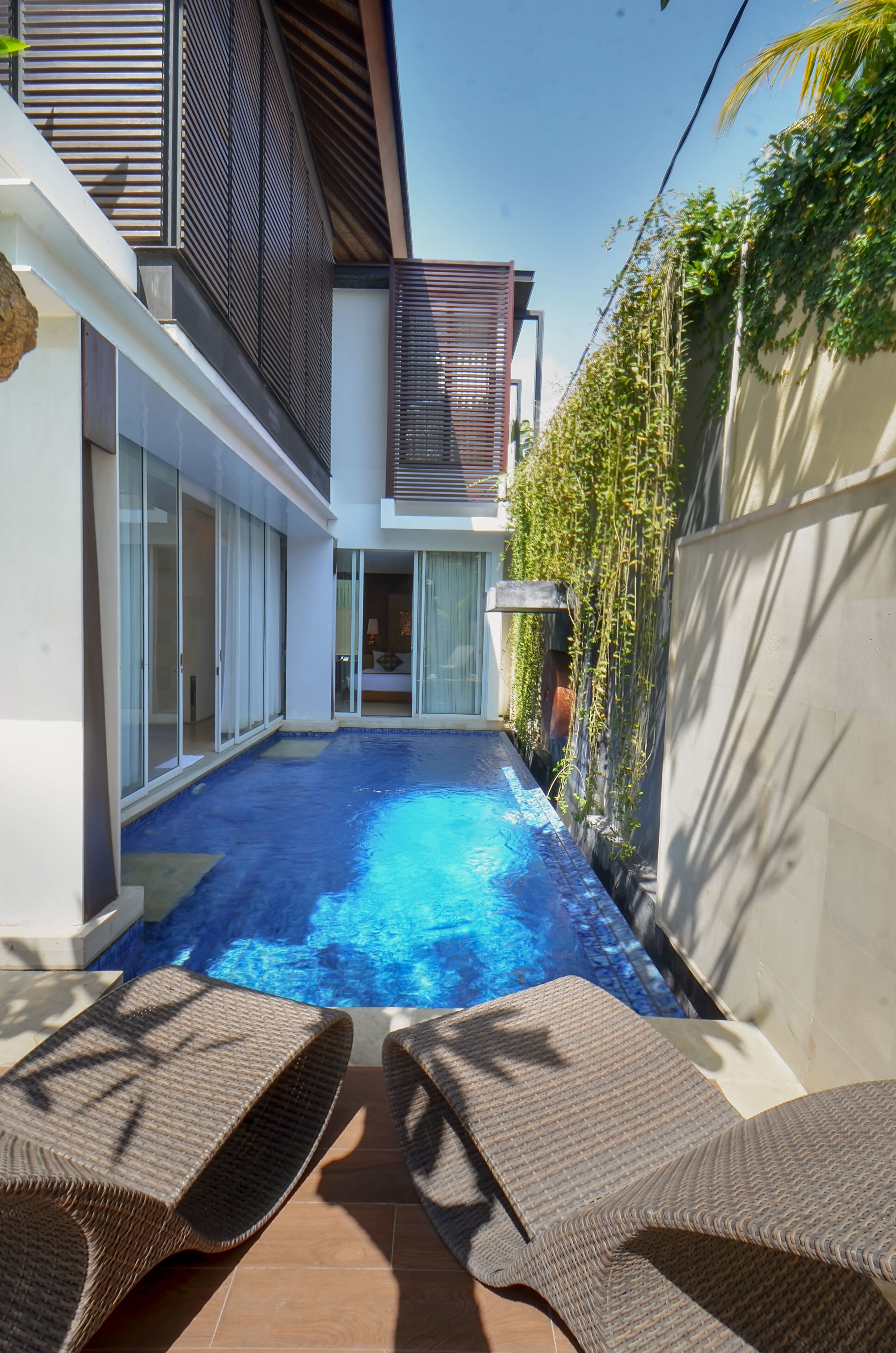 A modern outdoor pool area with a clear blue pool, two wicker lounge chairs, and a contemporary house with large glass doors and a balcony.
