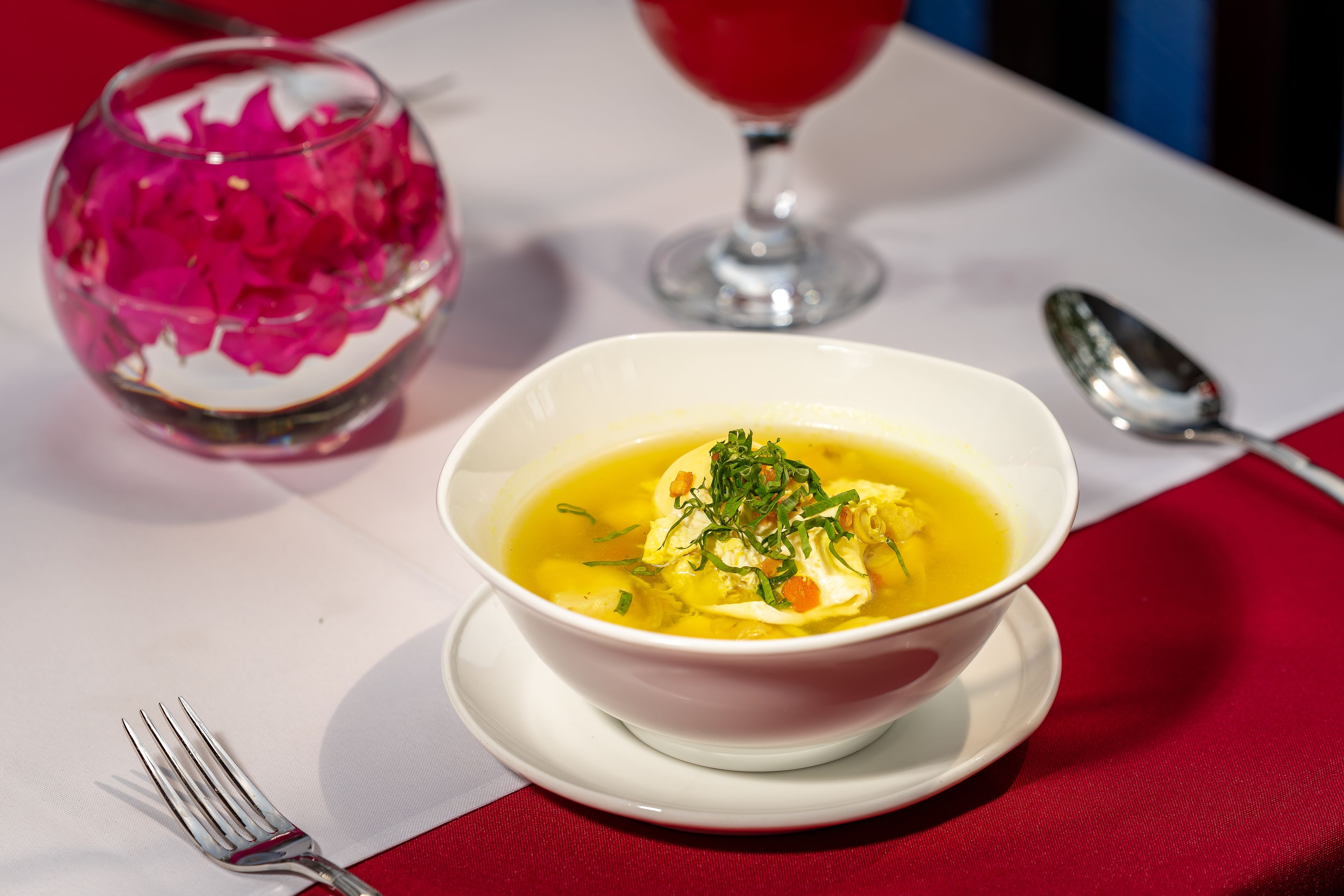 Un tazón de sopa con hierbas sobre un plato blanco, un vaso con flores rosadas y una cuchara sobre un mantel.