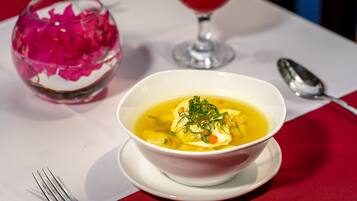 Un tazón de sopa con hierbas sobre un plato blanco, un vaso con flores rosadas y una cuchara sobre un mantel.