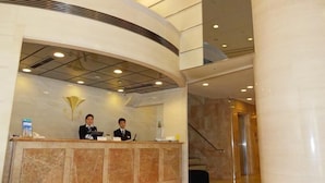 A reception desk with two staff members, a marble wall, and a staircase in the background. -  KOBE LUMINOUS HOTEL HYOGOEKIMAE (Kobe)