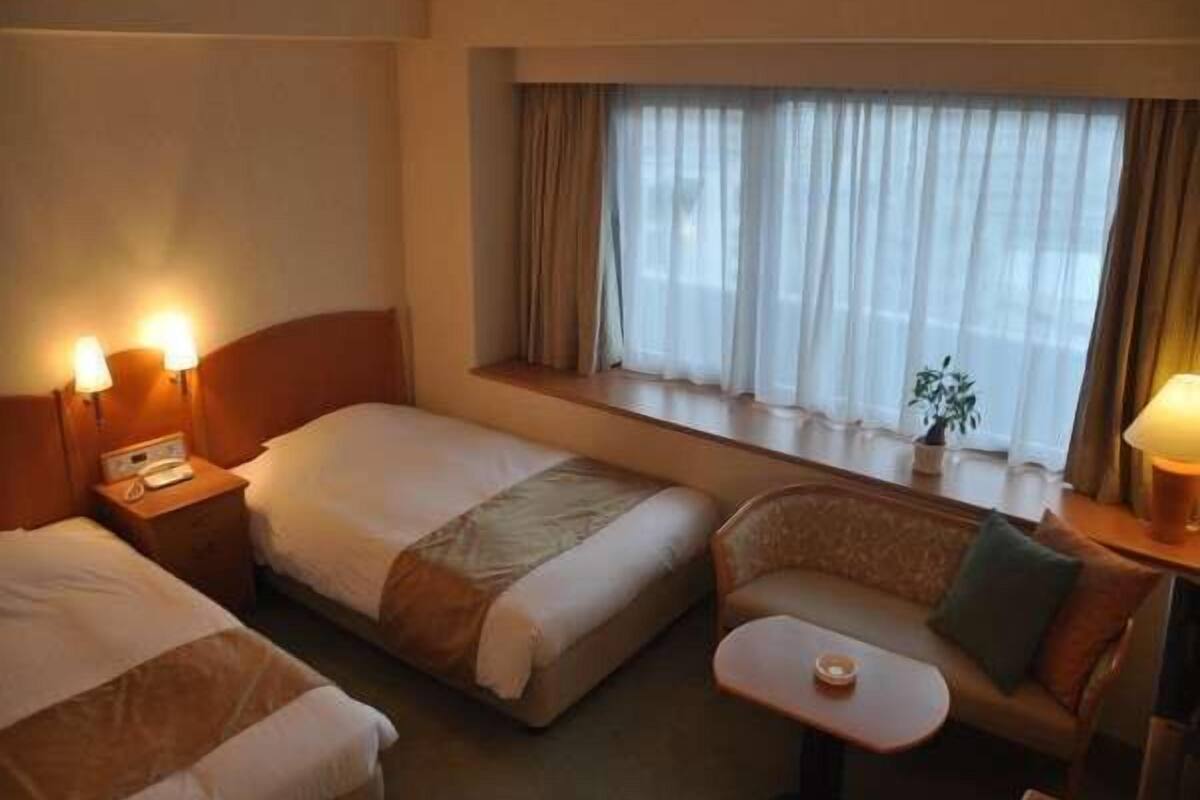 Habitación de hotel con dos camas, un pequeño sofá, una mesa y un ventanal con cortinas.