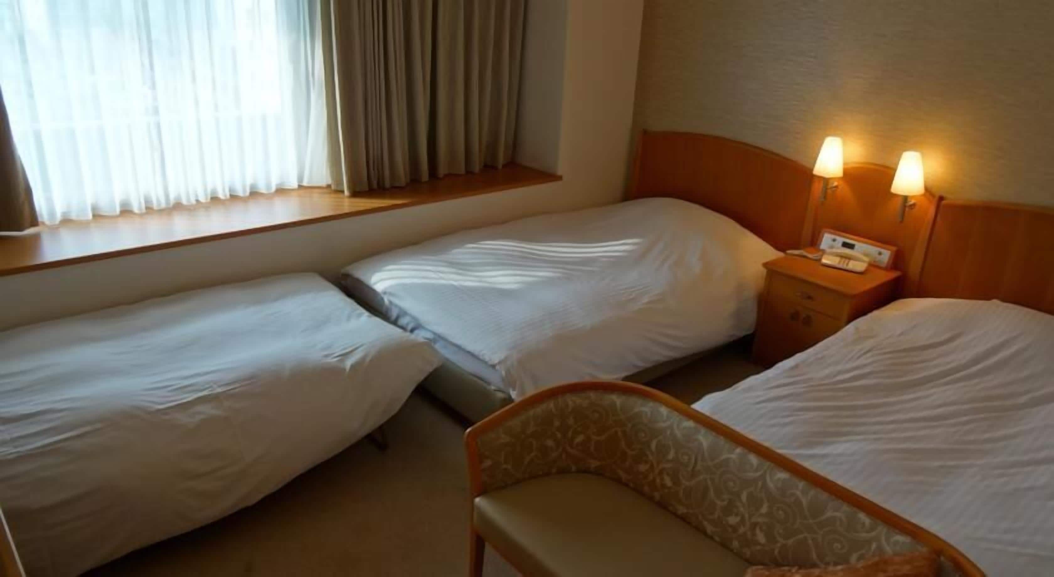 Una habitación de hotel con dos camas individuales, una mesita de noche de madera con lámpara y una ventana con cortinas.