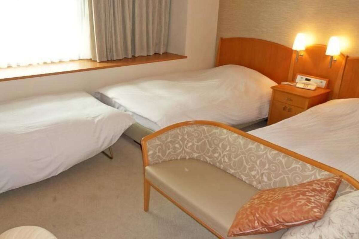 Habitación de hotel con dos camas individuales, un sofá cama, una mesita de noche con lámpara y una ventana con cortinas.
