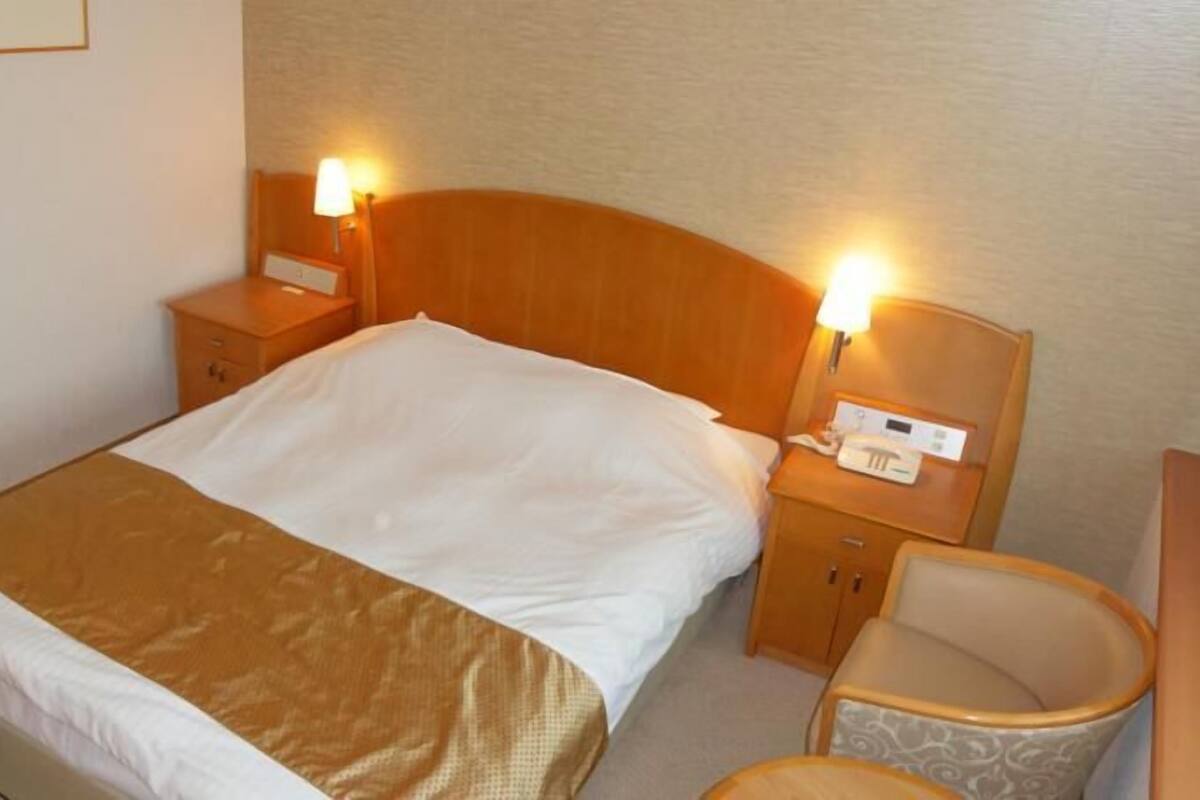 Habitación de hotel con una cama individual, una mesita de noche con lámpara, una silla y un pequeño escritorio con teléfono.