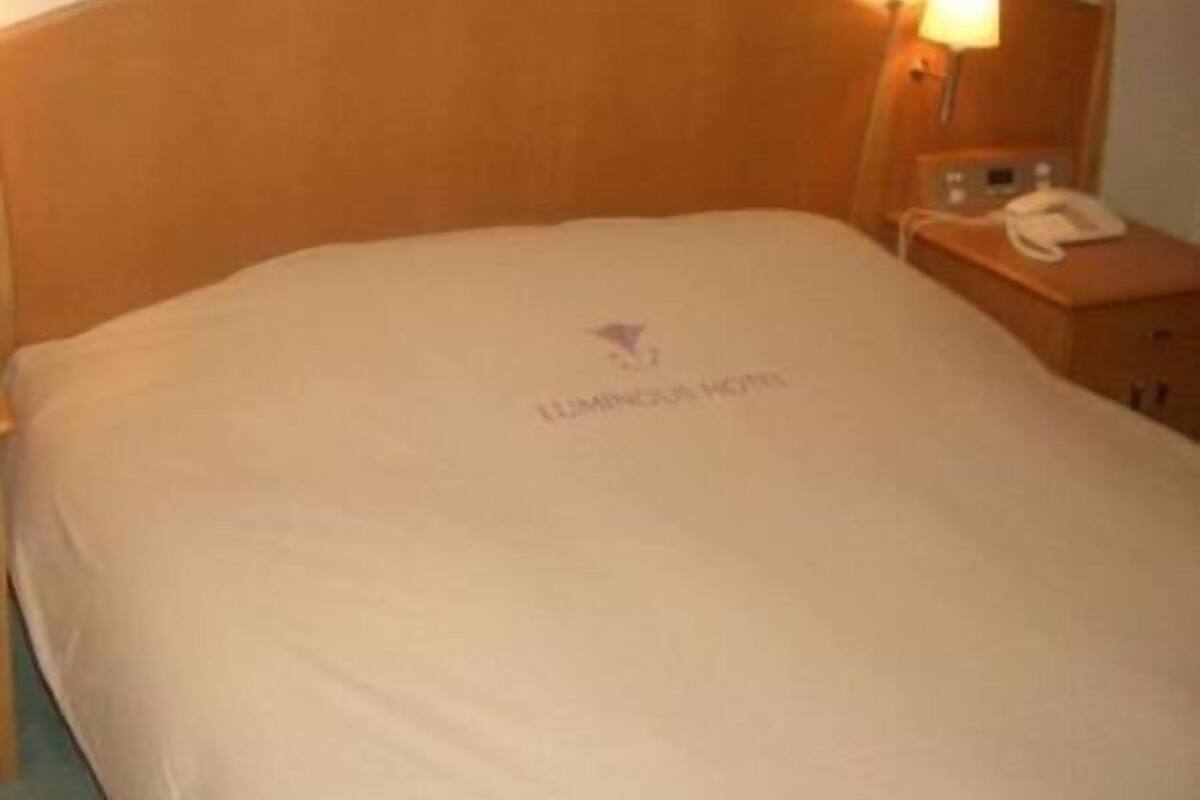 Habitación de hotel con cabecera de madera, mesita de noche con lámpara y cama con sábana blanca.