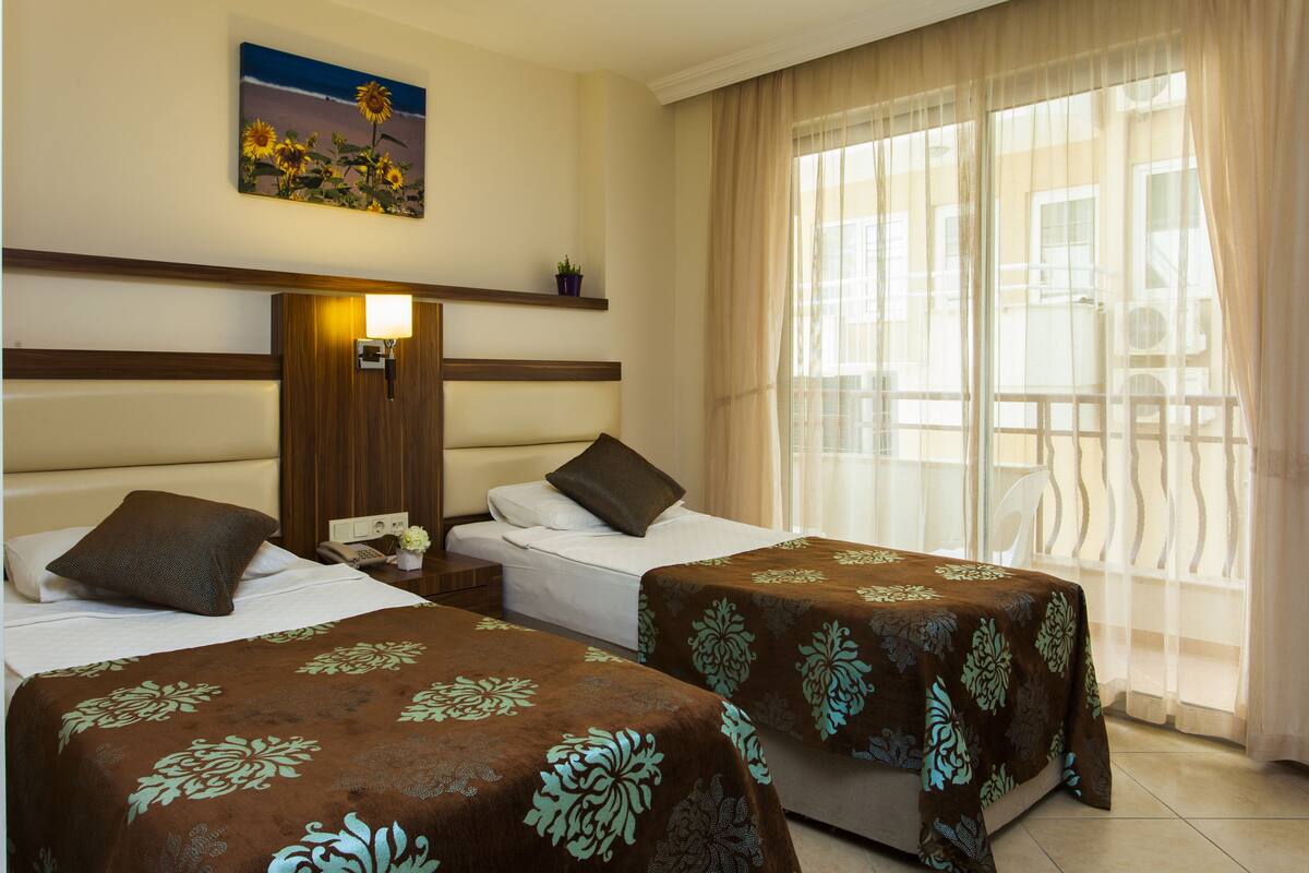 A hotel room with two beds, a nightstand with a lamp, a painting of sunflowers, and a window with sheer curtains.