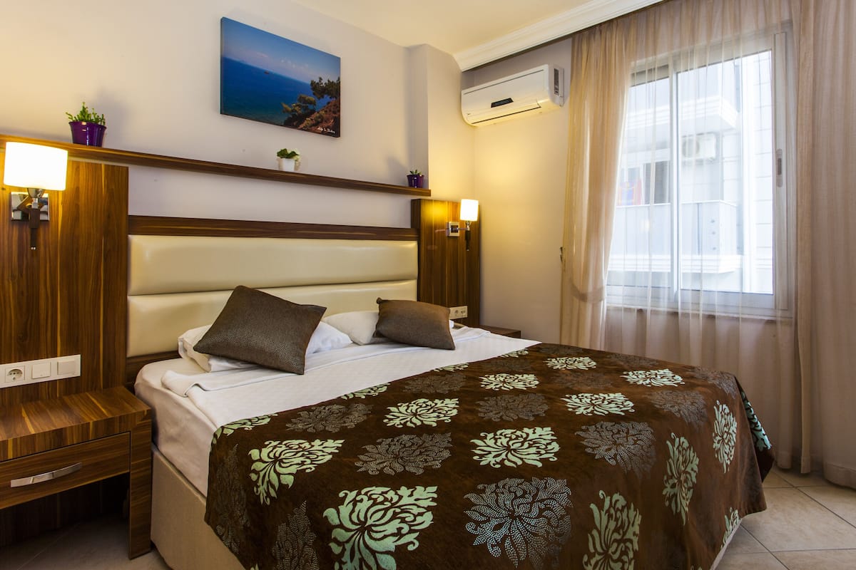 A hotel room with a bed, bedside tables, a nightstand, a wall-mounted air conditioner, a window with curtains, and a painting on the wall.