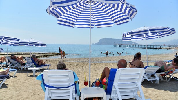 Una spiaggia con lettini bianchi e ombrelloni a righe blu, un molo sullo sfondo e persone che nuotano nell'acqua.