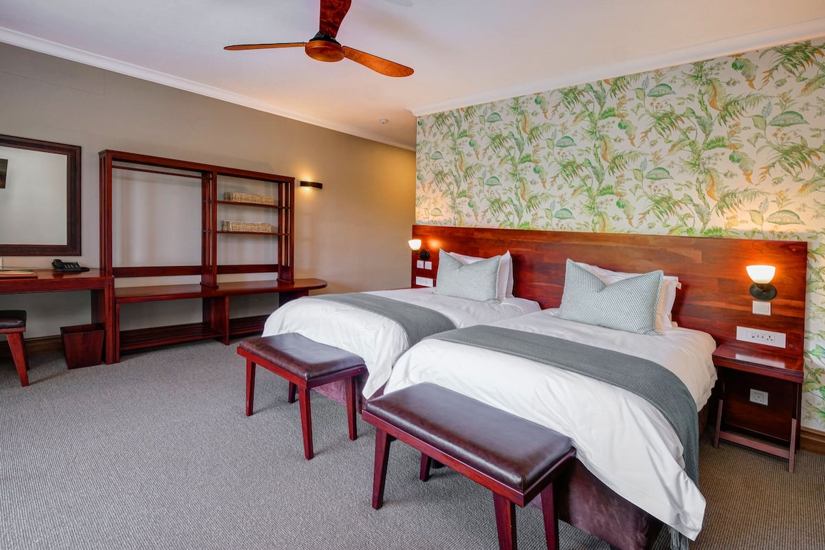 A hotel room with two beds, a wooden desk, a bench, and a ceiling fan.