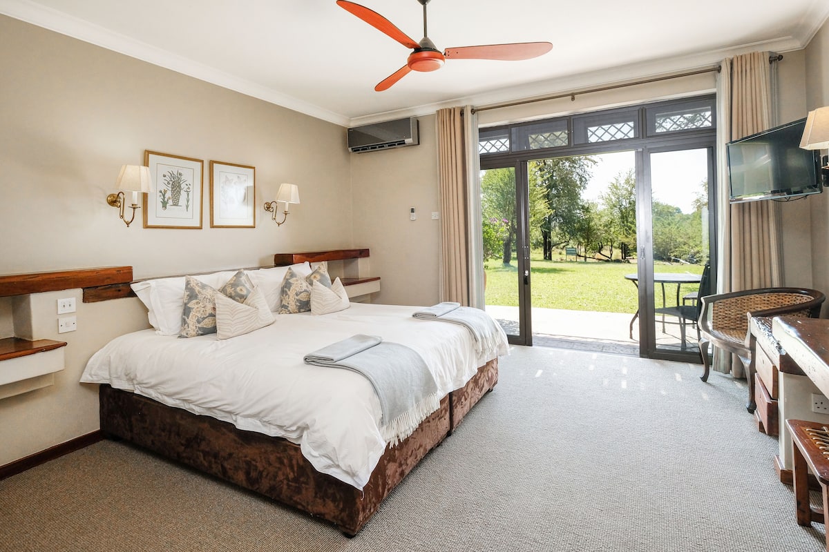 A hotel room with a large bed, a desk, a TV, and a view of the garden.