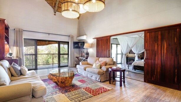 A spacious living area with a sofa, a coffee table, and a large window offering a view of the outdoors. - Ilala Lodge Hotel (Victoria Falls)