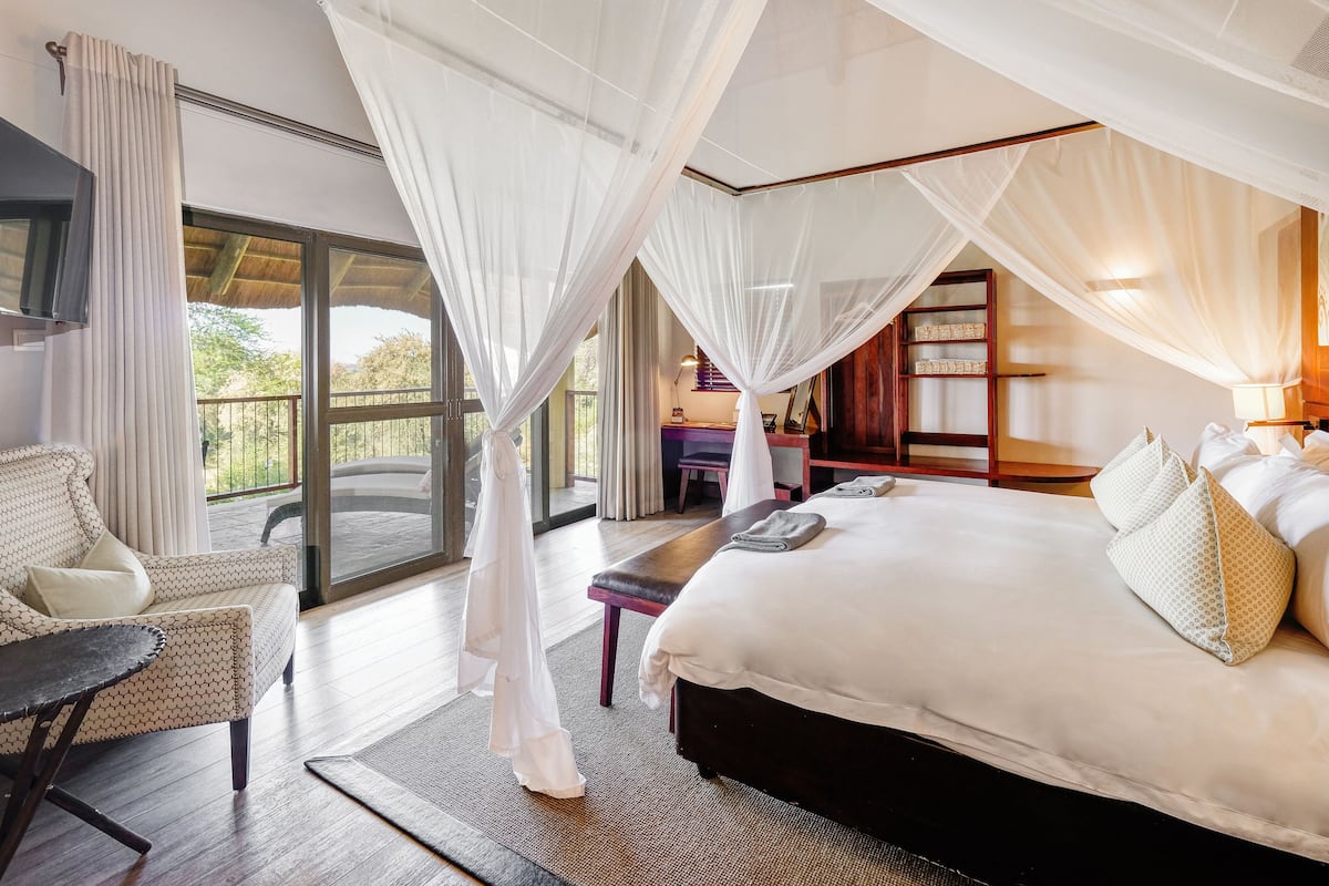 A bedroom with a large bed, a chair, a desk, and a view of the outdoors.