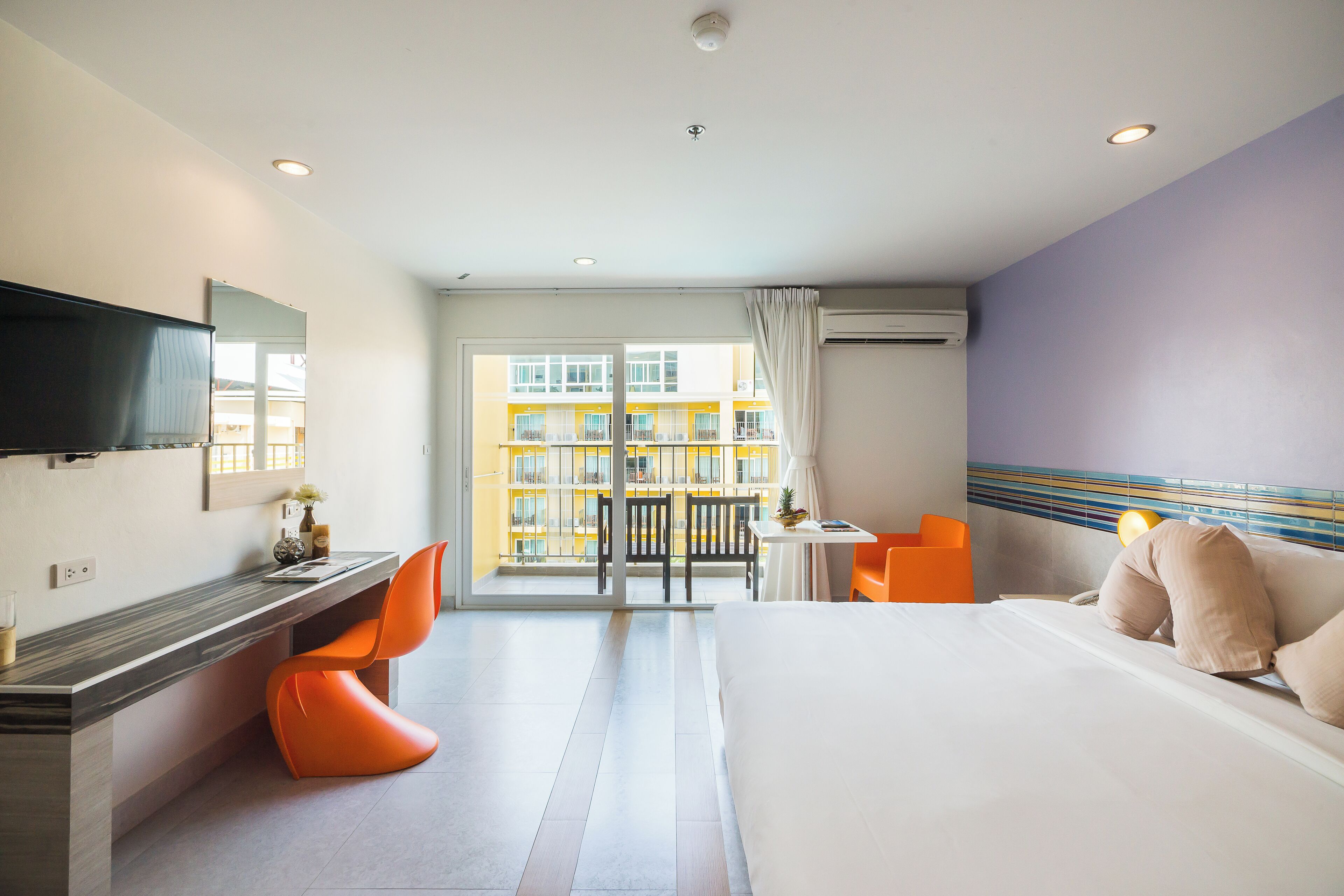 A hotel room with a bed, a desk with a chair, a TV, and a view of a building through the window.