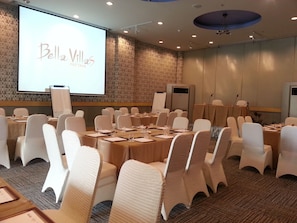Ein Konferenzraum mit runden Tischen, weißen Stühlen und einer großen Leinwand, auf der 'Bella Villa Pattaya' zu sehen ist.