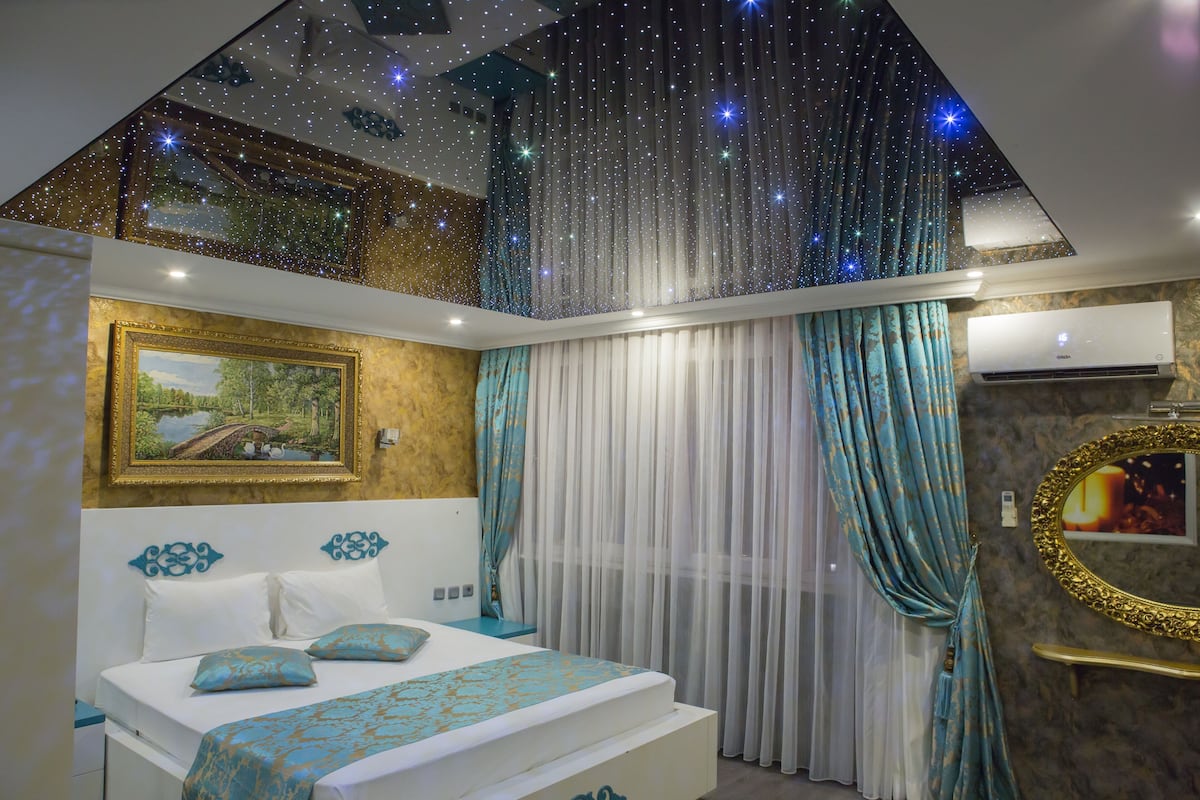 A bedroom with a bed, curtains, a painting, and a ceiling with star-like lights.