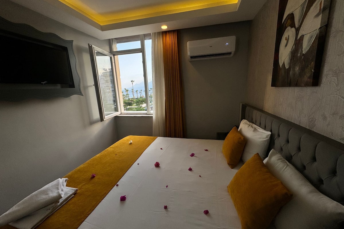 A hotel room with a bed, a flat-screen TV mounted on the wall, an air conditioning unit, and a window with a view of palm trees and mountains.