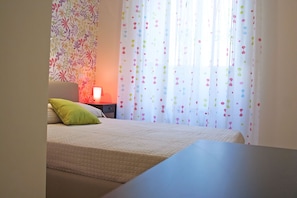 A neatly made bed with a green pillow, a bedside table with a lamp, and a window with colorful curtains. - City-In Hostel B&B (Catania)