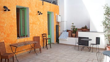 Una veranda esterna con tavolo e sedie, un piccolo giardino con piante in vaso e un muro bianco con una finestra e persiane verdi.