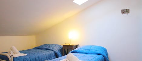 Een kleine, eenkameraccommodatie met twee bedden, elk met een blauwe dekbedovertrek en witte kussens. Er is een nachtkastje met een lamp tussen de bedden.