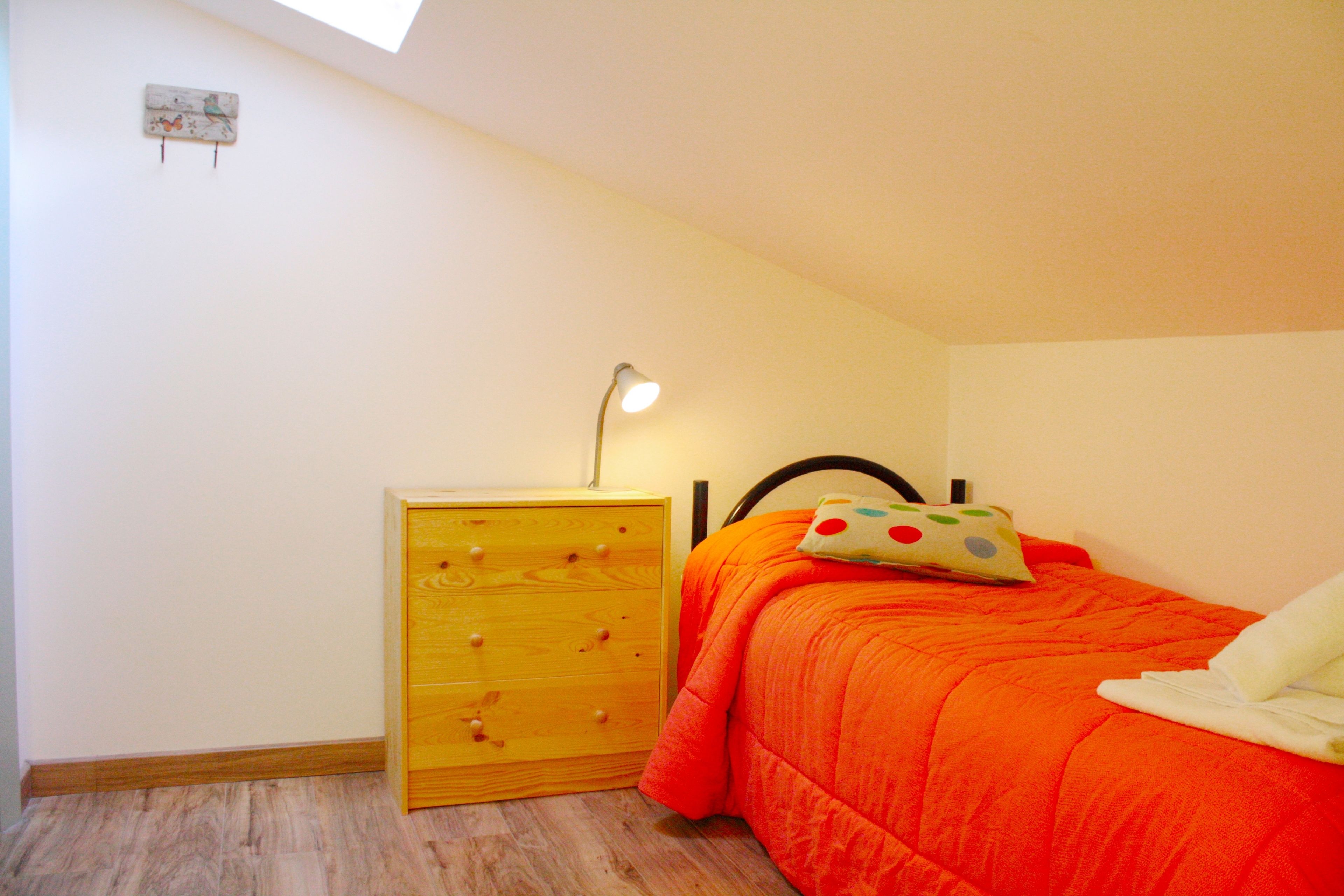 A bedroom with a wooden dresser, a bed with an orange bedspread, a nightstand with a lamp, and a small wall decoration.