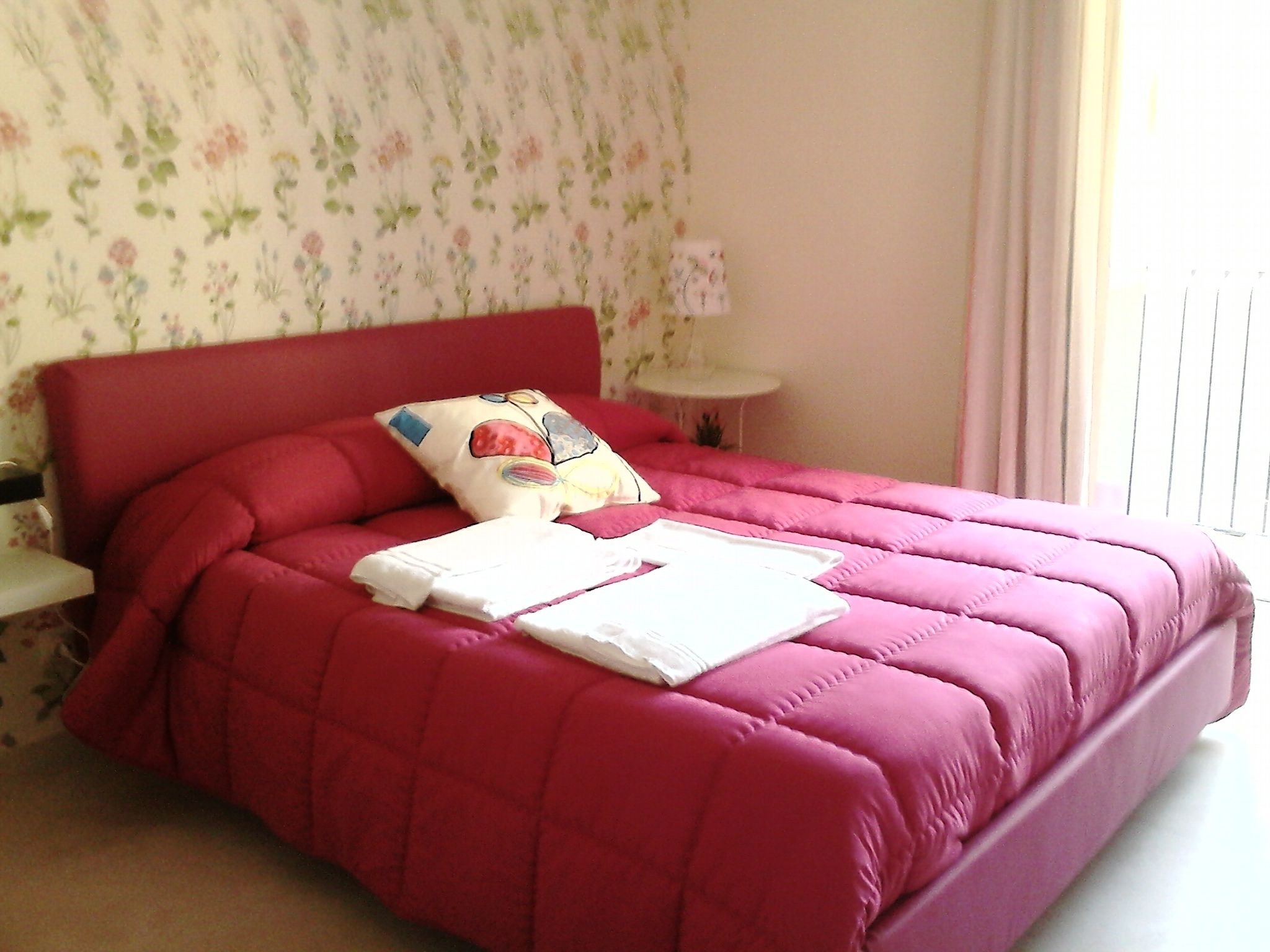 A neatly made bed with a red headboard and a floral-patterned wall.
