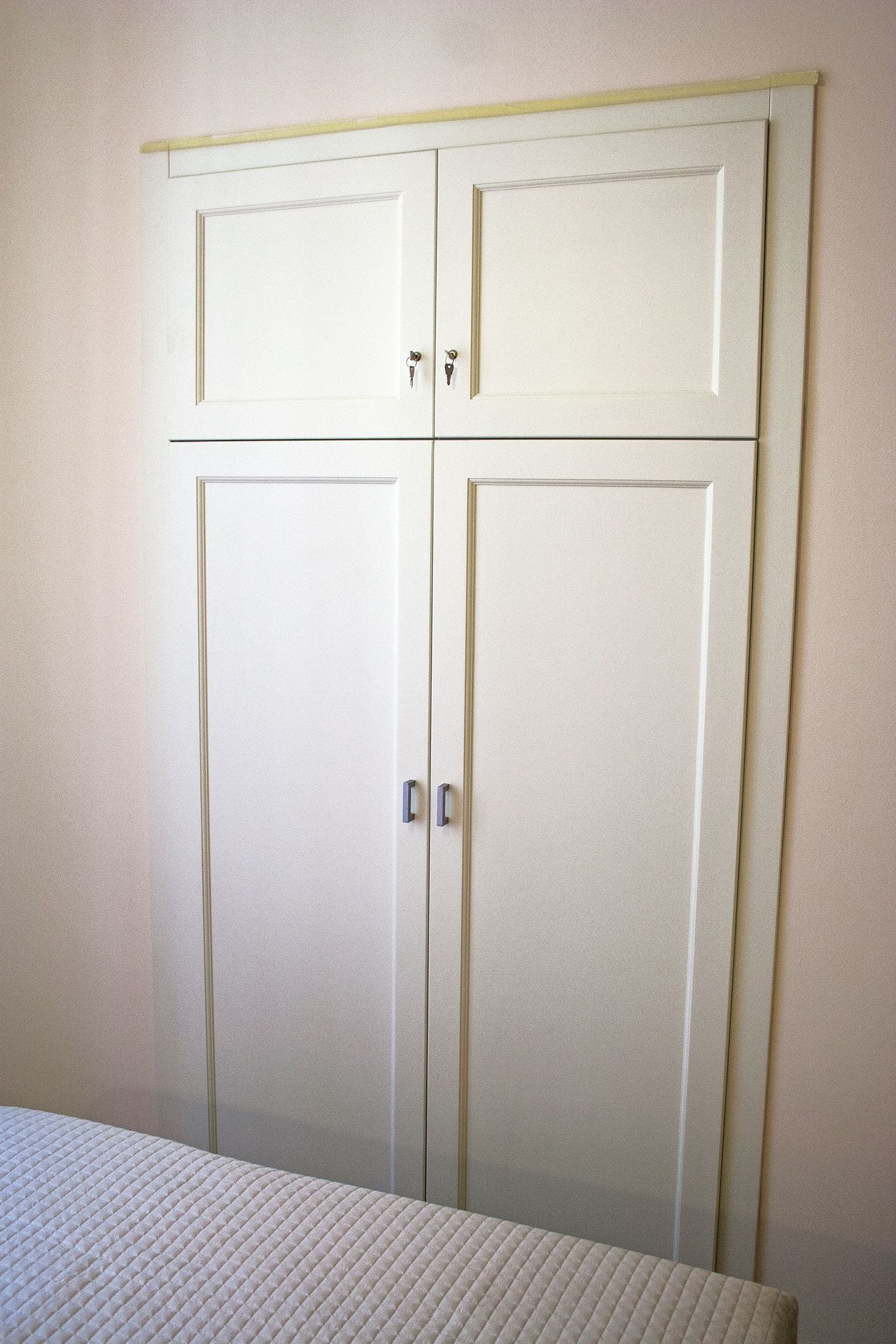 Ein weißer Kleiderschrank mit zwei Türen und einer kleinen Ablage, ein Bett mit gesteppter Decke und eine schlichte Wand.
