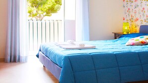 A neatly made bed with a blue bedspread and white towels, a window with curtains, and a nightstand with a lamp.