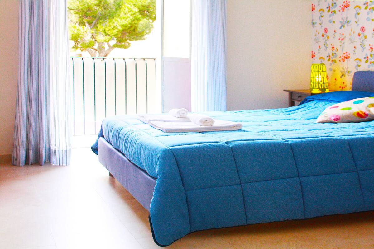 A neatly made bed with a blue bedspread and white towels, a window with curtains, and a nightstand with a lamp.