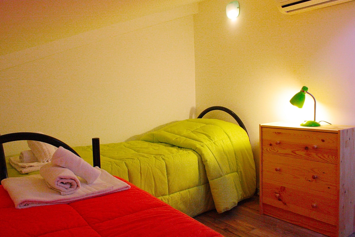 A single bed with a red blanket, a green bedspread, and a wooden nightstand with a green lamp.