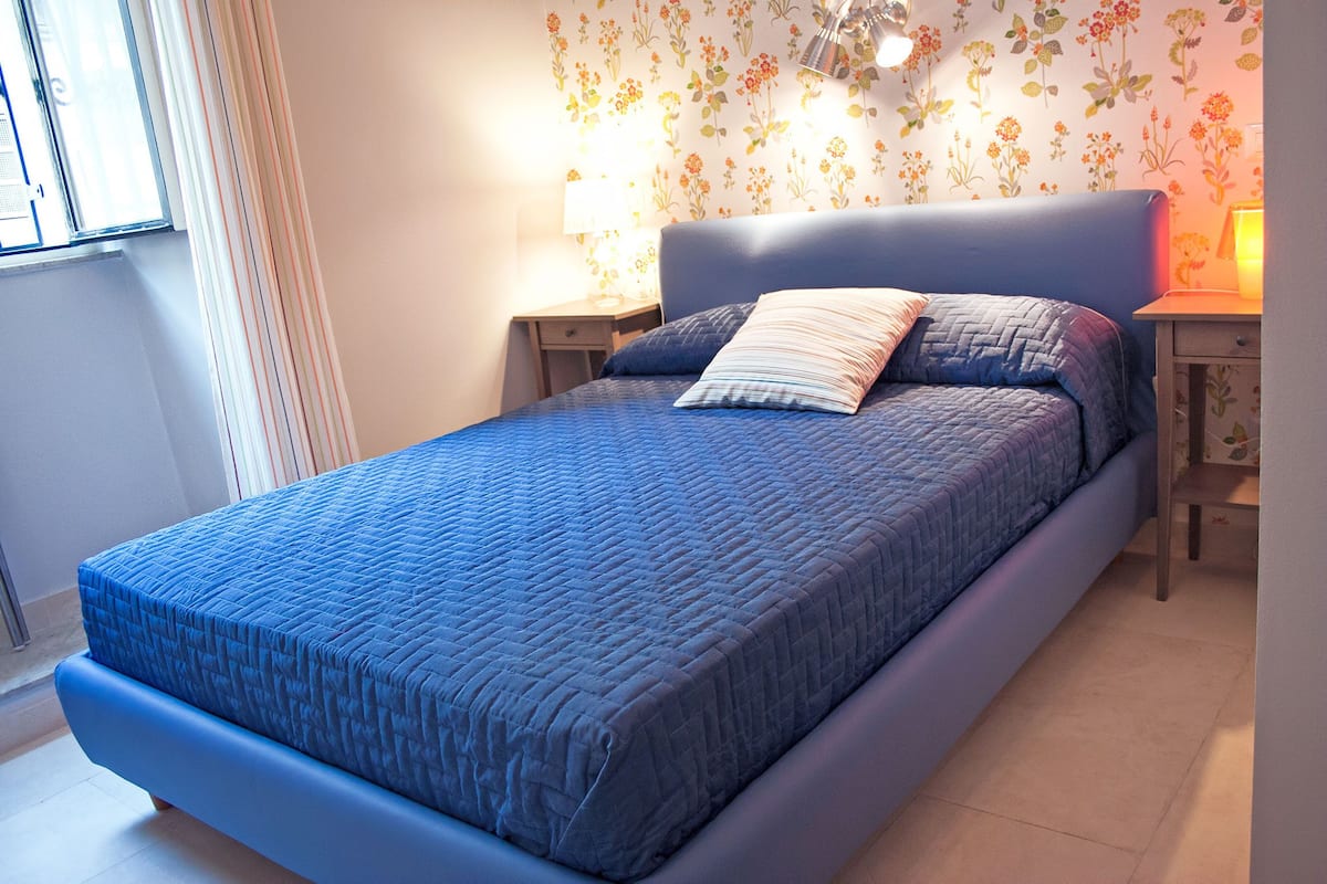 A bedroom with a bed, two bedside tables, a window, and floral wallpaper.