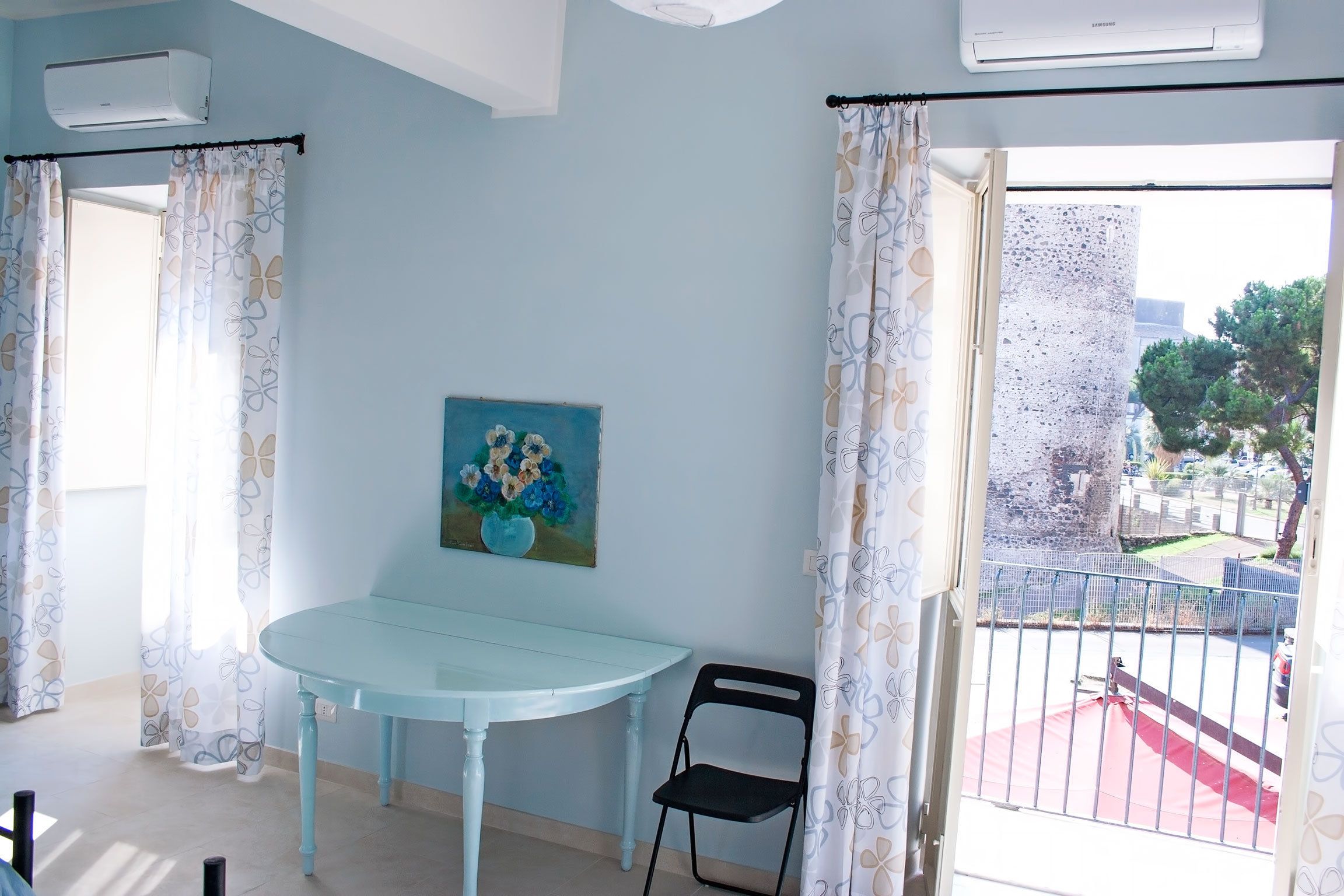 A room with a light blue table, a black chair, a painting on the wall, and a view of a courtyard through a window with patterned curtains.