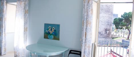 A room with a light blue table, a black chair, a painting on the wall, and a view of a courtyard through a window with patterned curtains.