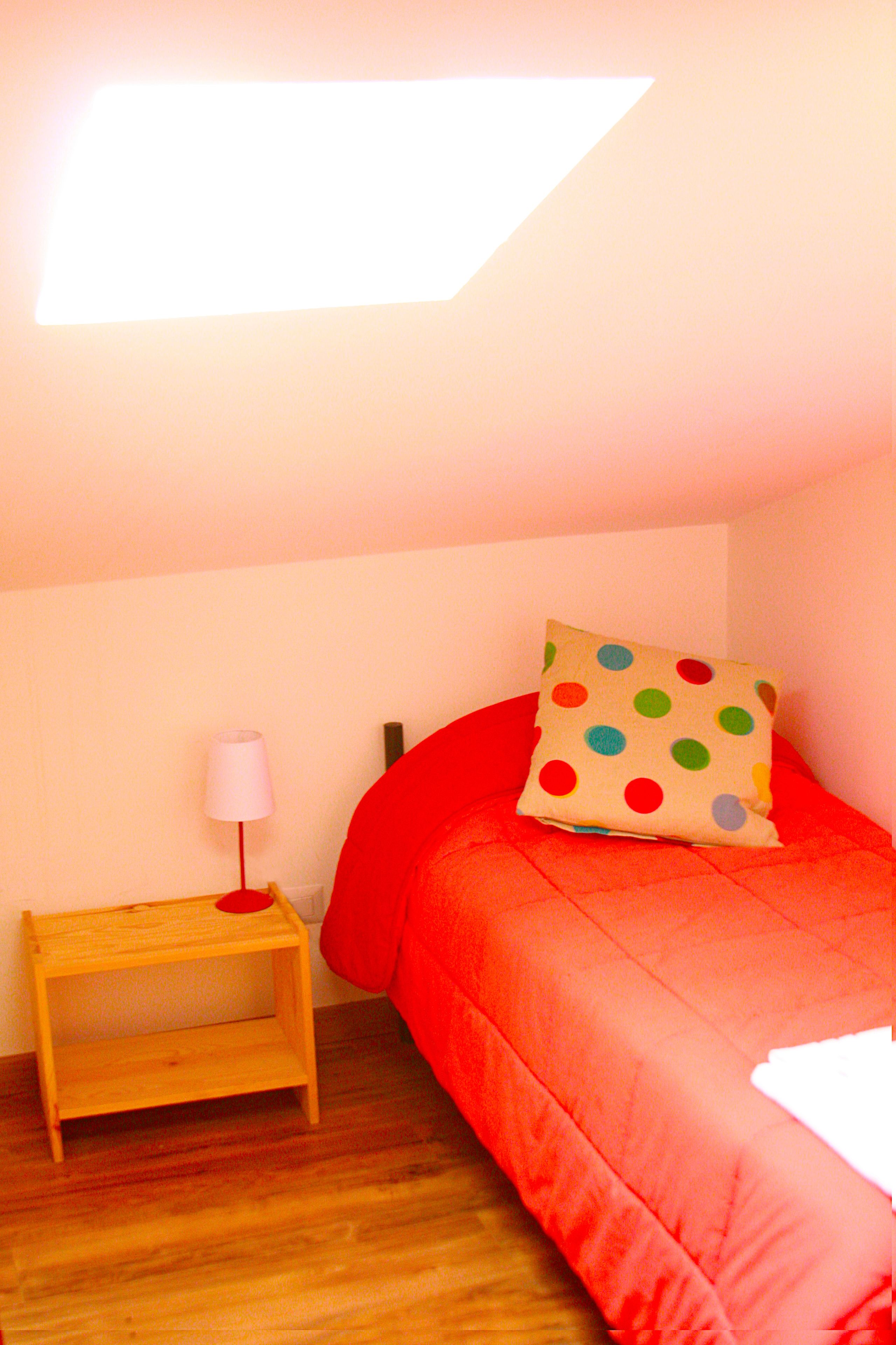 Un dormitorio con una cama roja, un almohadón de lunares, una mesita de noche de madera y una lámpara, y un tragaluz arriba.