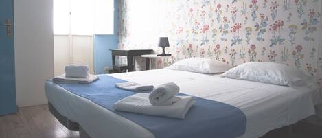 Cama bem arrumada com lençóis brancos e um toque de azul, uma pequena mesa com abajur e um papel de parede estampado com flores.