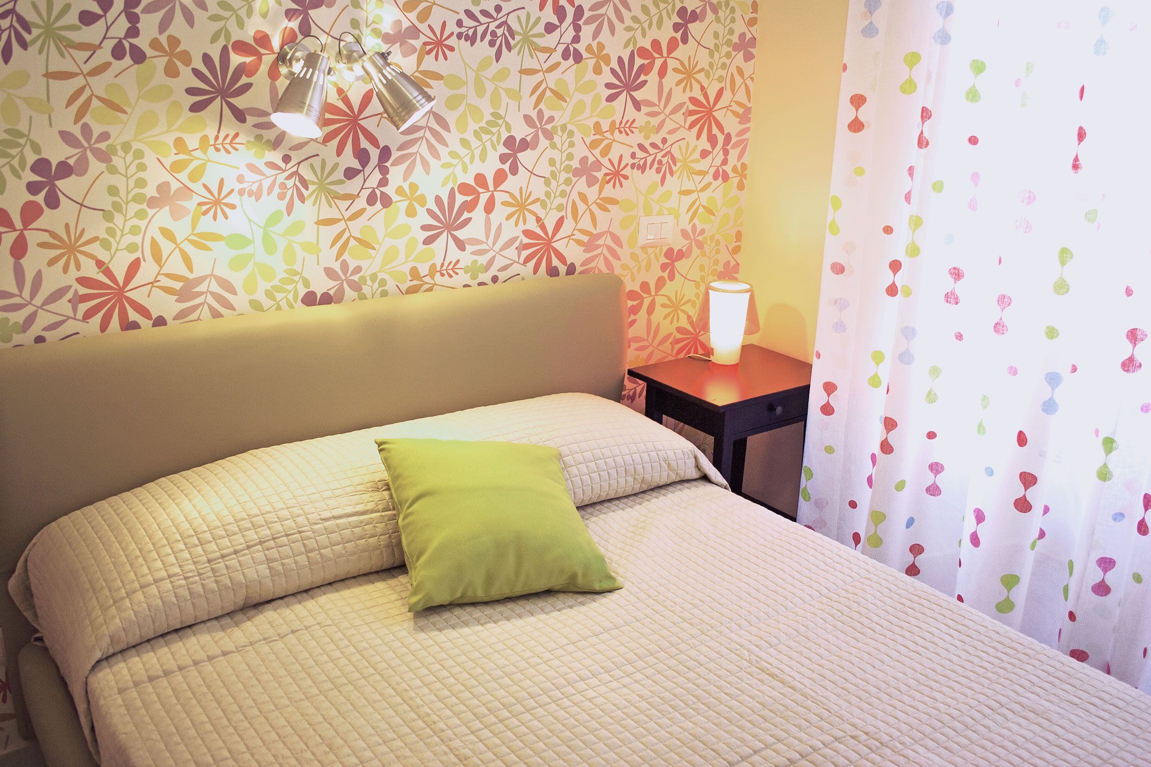 Een net opgemaakt bed met een gewatteerde spreidek en twee kussens, een groen en een wit, in een kamer met bloemenbehang en een klein nachtkastje met een lamp.