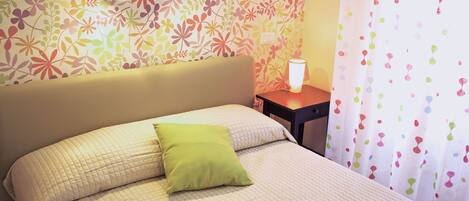 A neatly made bed with a quilted bedspread and two pillows, one green and one white, in a room with floral wallpaper and a small bedside table with a lamp.