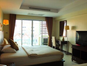 A hotel room with a bed, a desk, a television, and a view of buildings through the window. - August Suites (Pattaya)