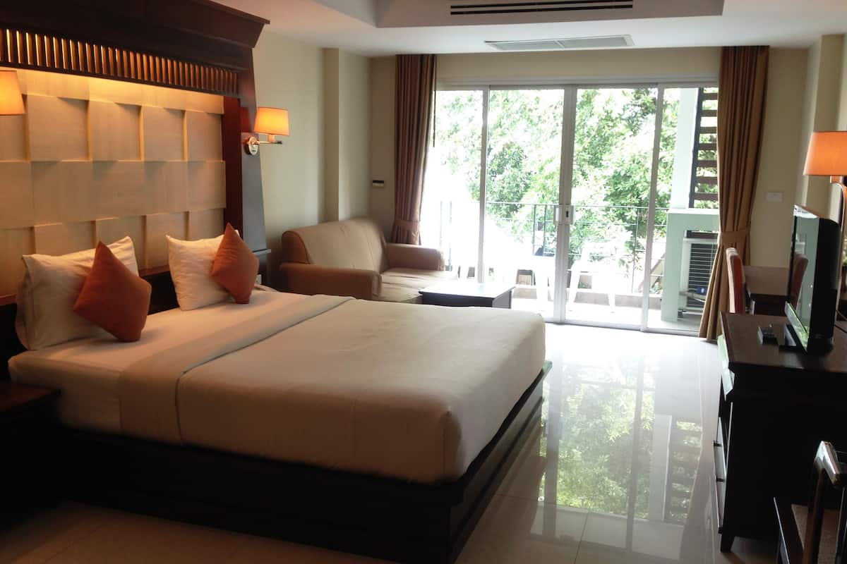 A hotel room with a large bed, two armchairs, a desk with a TV, and a balcony with a view of greenery.