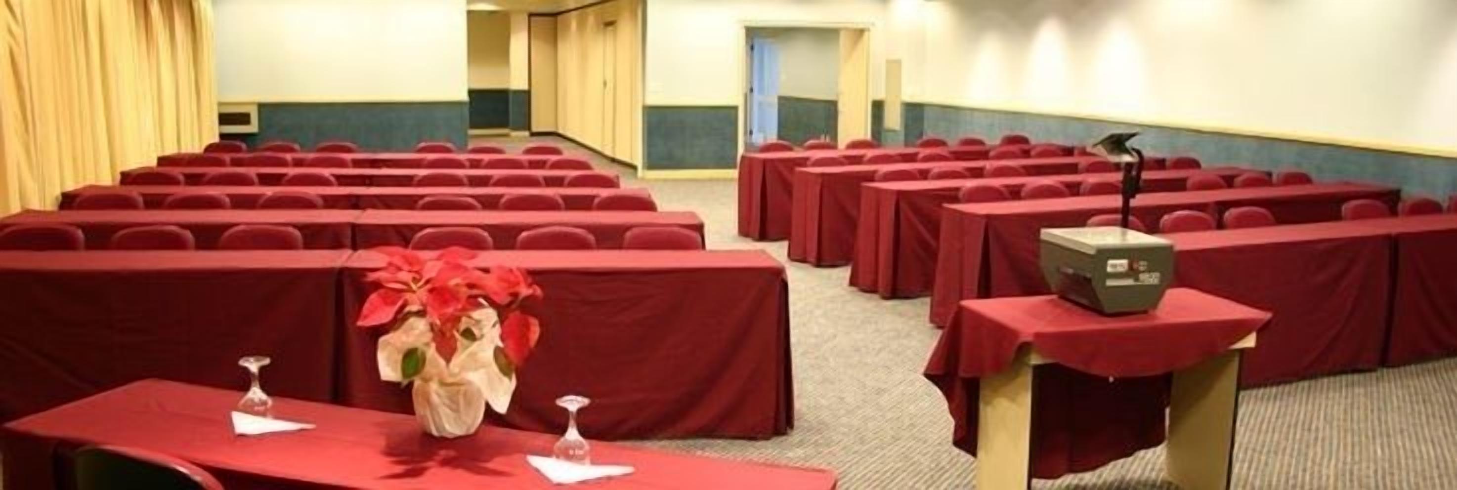 Konferenssisali, jossa on riveissä pöytiä, jokainen peitetty punaisella pöytäliinalla, ja puhujapönttö mikrofonin kanssa.