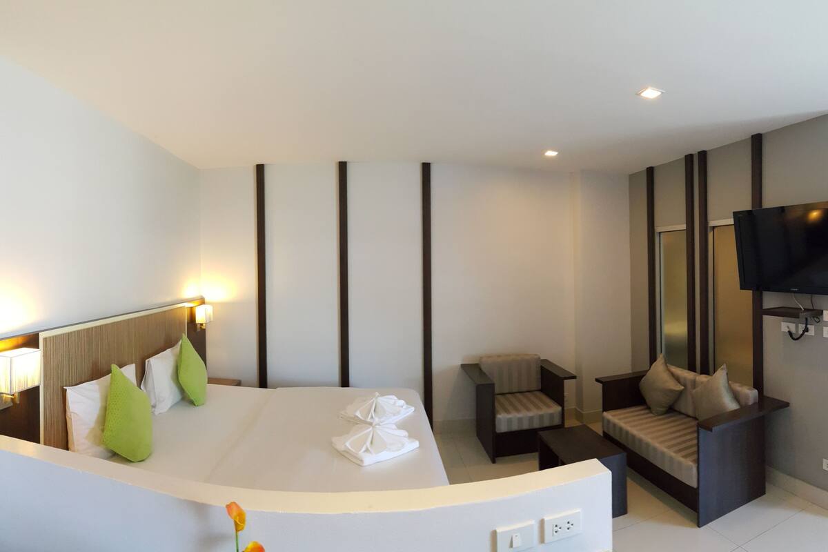Une chambre d’hôtel moderne dotée d’un grand lit, d’un coin salon avec un canapé et un fauteuil, d’un téléviseur à écran plat fixé au mur et d’une petite table avec une lampe.