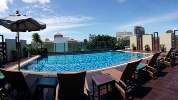 A rooftop pool with clear blue tiles, surrounded by wicker lounge chairs and a large umbrella.