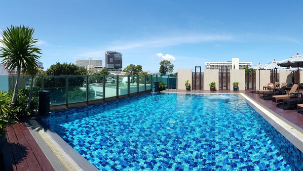 A rooftop pool with clear blue water, surrounded by lounge chairs and umbrellas, with city buildings in the background.