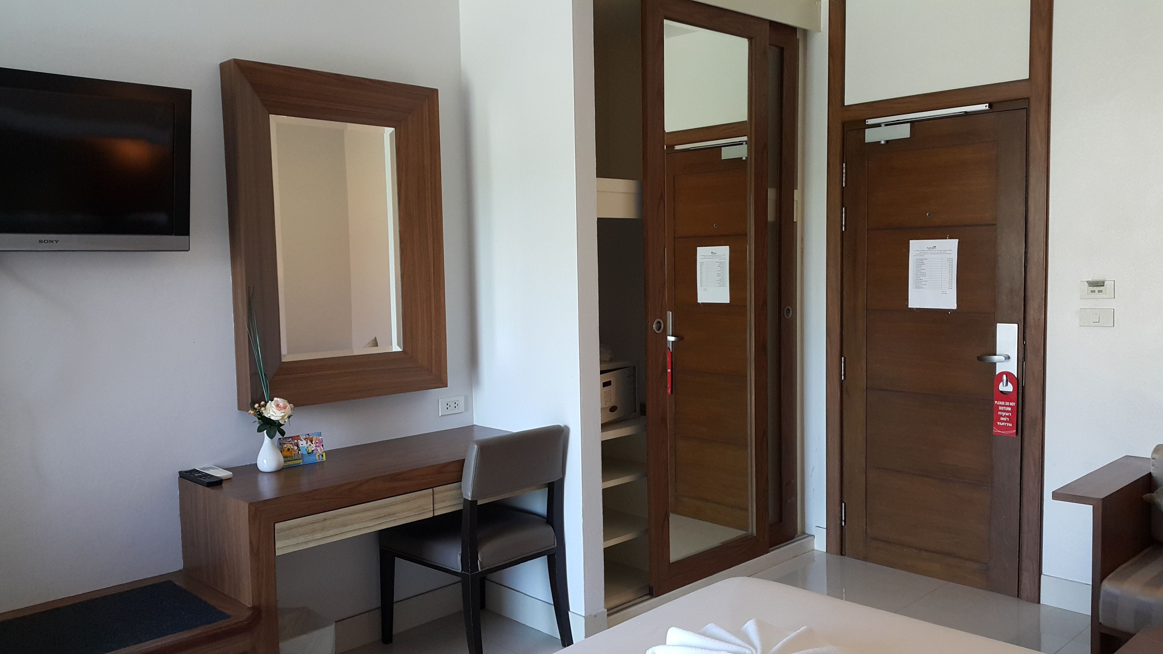 A hotel room with a wooden desk, a chair, a TV mounted on the wall, and a large mirror.