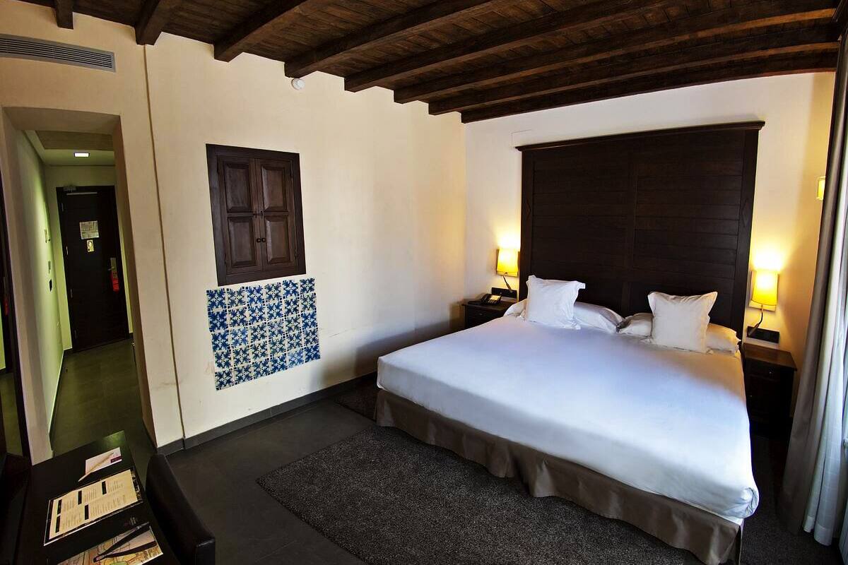 A hotel room with a large bed, two bedside tables with lamps, a dark wooden headboard, a patterned wall decoration, and a door leading to another room.