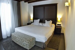 A hotel room with a large bed, a bedside table with a lamp, a bench, and a headboard with a grid pattern.