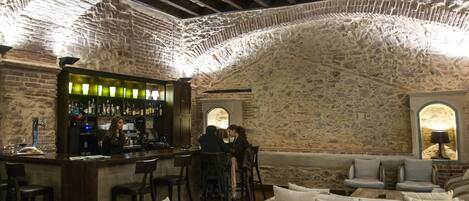 Una zona de bar con paredes de piedra, vigas de madera y una barra con taburetes. Hay áreas de estar con cojines y una lámpara en la esquina.