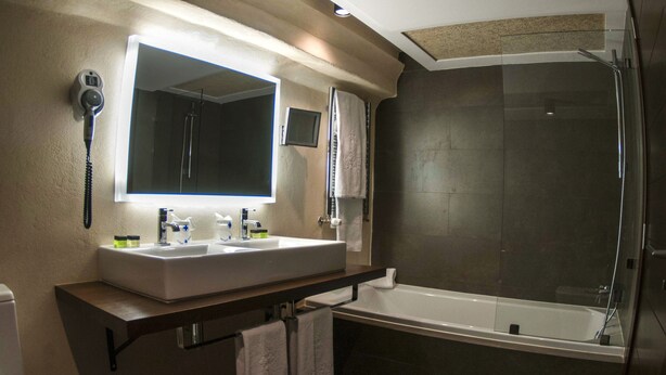 A modern bathroom with a dual sink vanity, a large mirror, a glass-enclosed shower, and a built-in toilet. - Hotel Convento Aracena & Spa (Aracena)