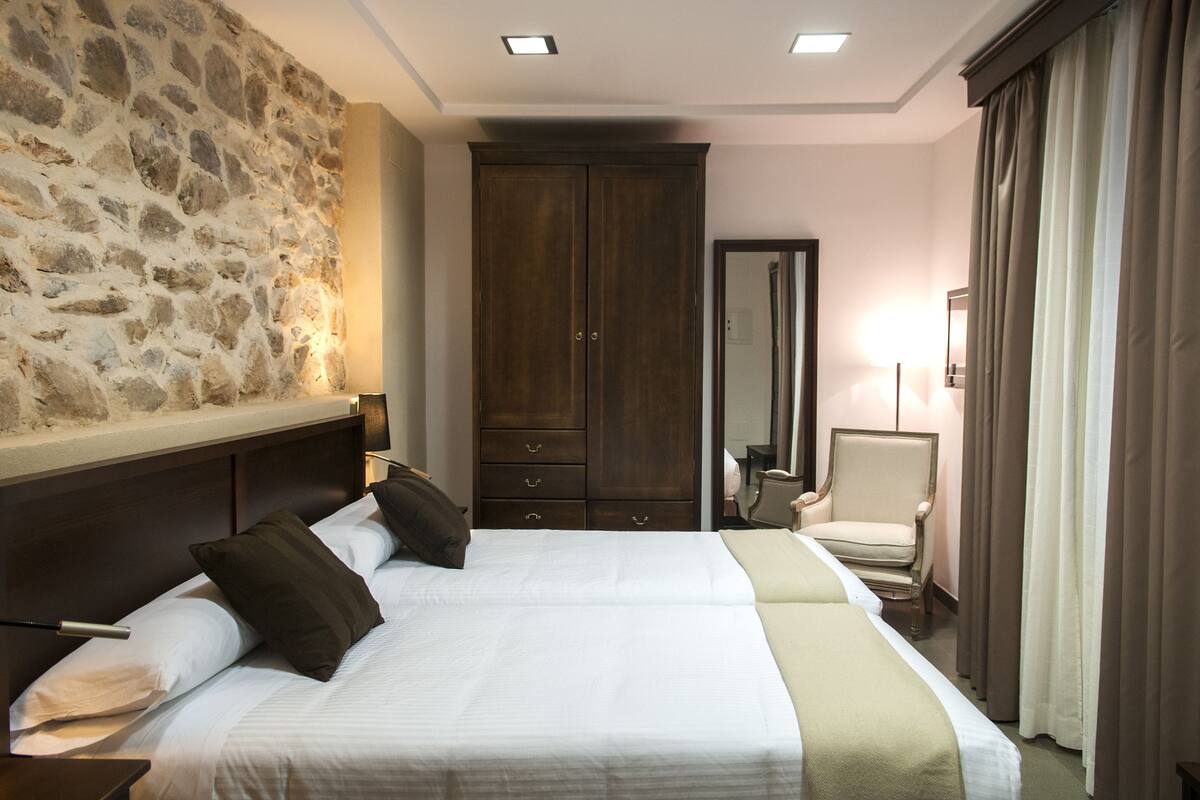 A hotel room with a stone accent wall, a large wardrobe, a bed with white linens, and a chair.