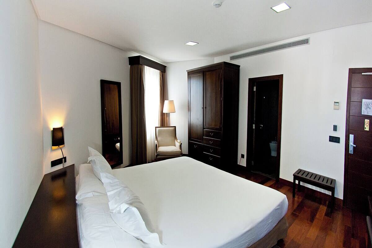 A hotel room with a large bed, a chair, a desk, and a wardrobe.