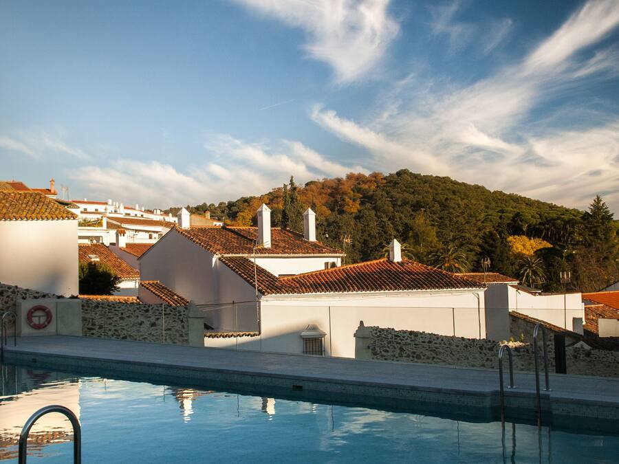 A swimming pool with a stone wall and metal railings, surrounded by buildings with red-tiled roofs and a clear sky.